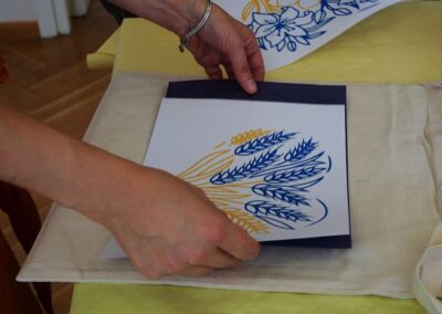 Dłonie kobiety kladące na stole kartkę z namalowanym żółto-niebieskim wzorem kwiatowym.